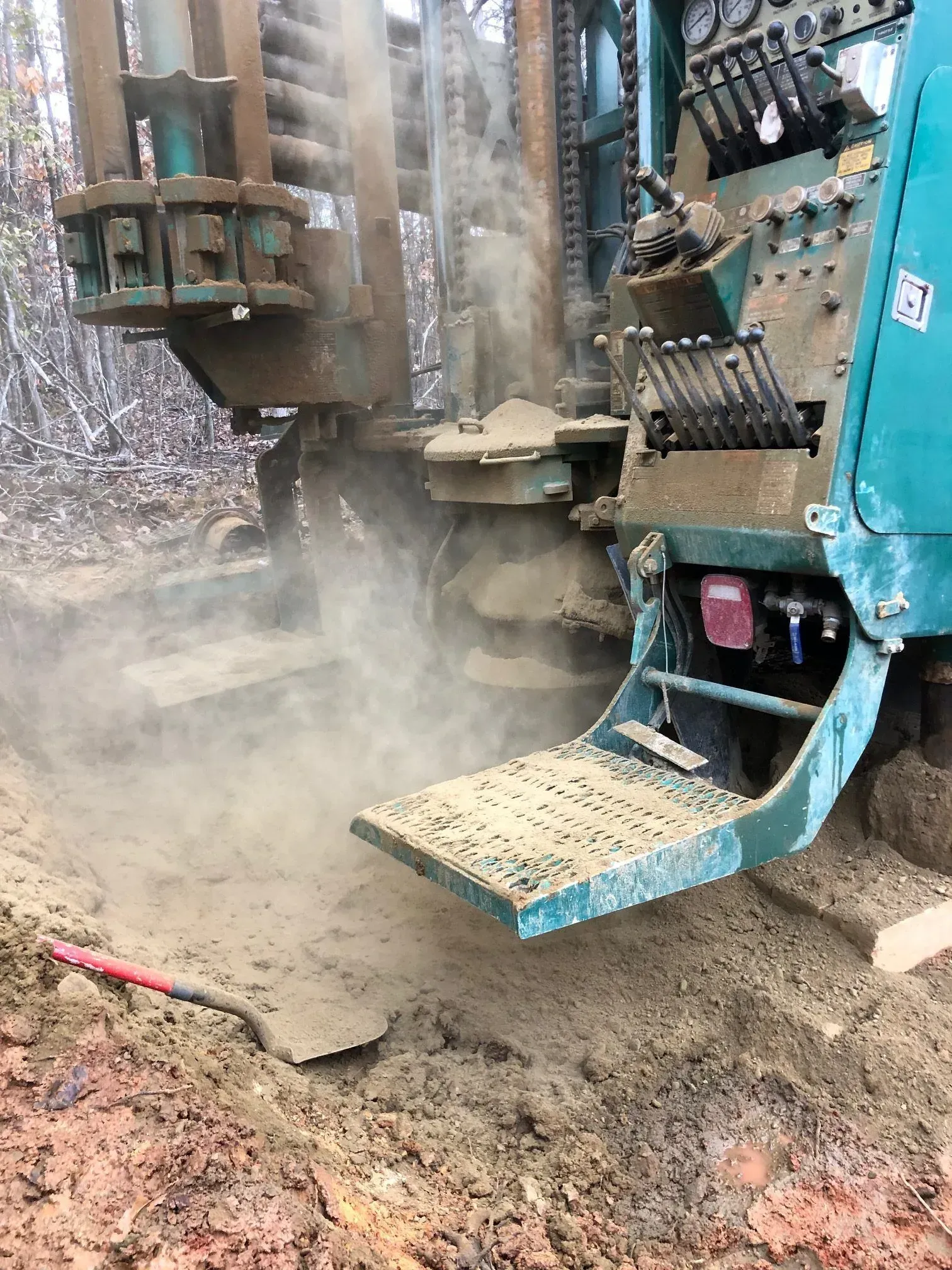 Drilling rig in operation, raising dust. Turquoise machinery drills into muddy ground outdoors.