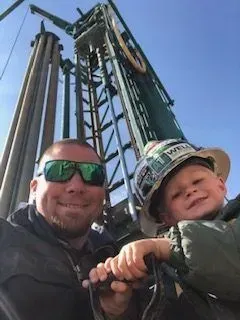 Man and child smiling, posed in front of oil derrick. Child wearing a hard hat, outside.