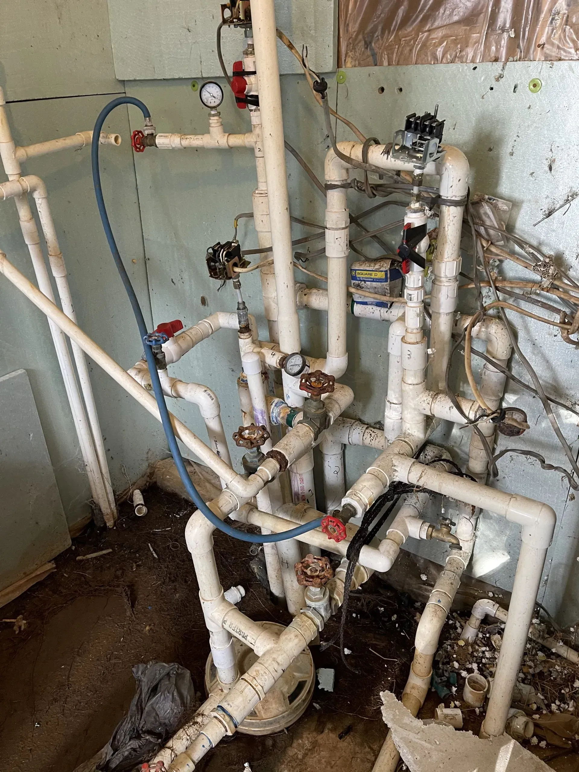White plumbing pipes, valves, and gauges in a utility room; blue hose.