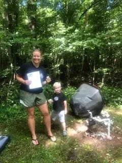 Woman and child stand near a large rock formation in a wooded area. The woman holds a paper and smiles.