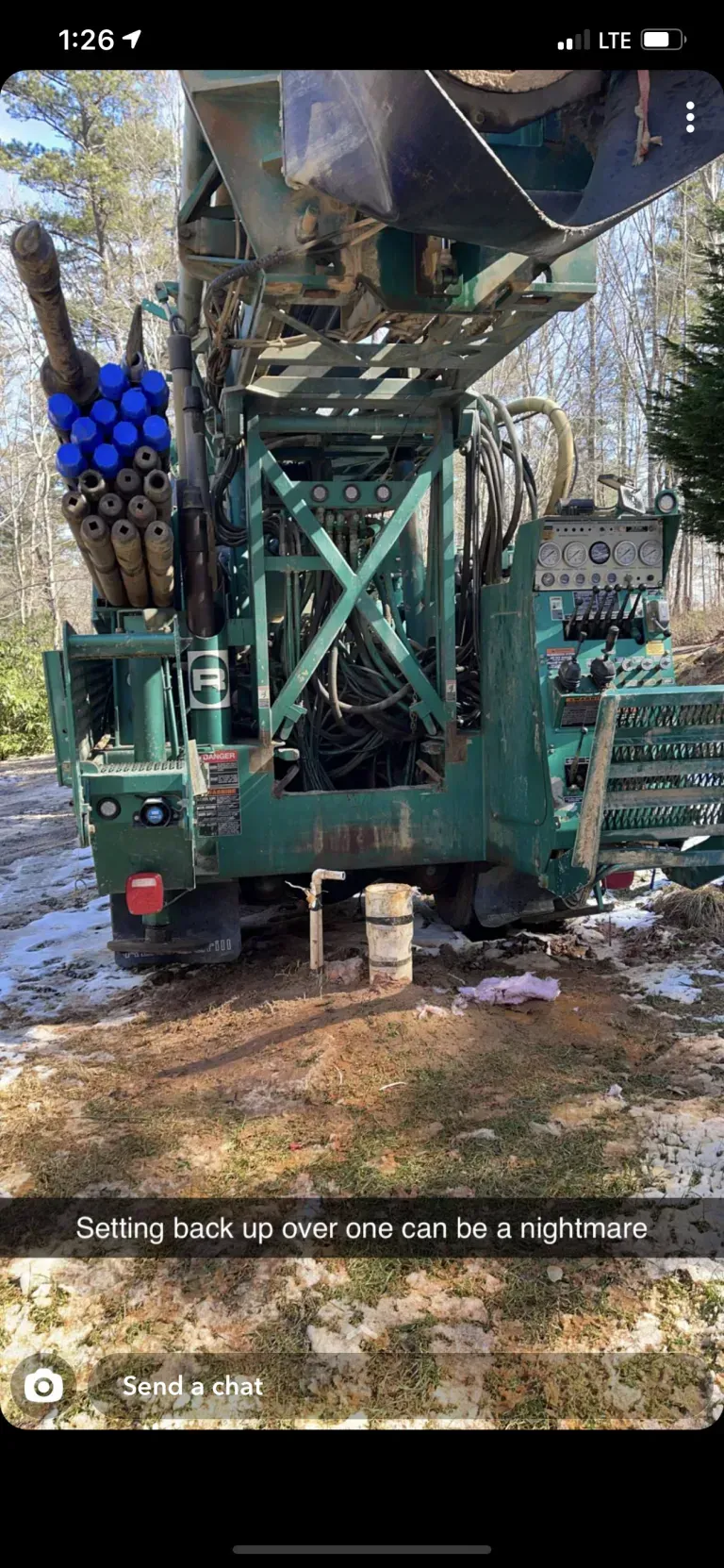 A large green drilling rig in a wooded area, with snow on the ground.