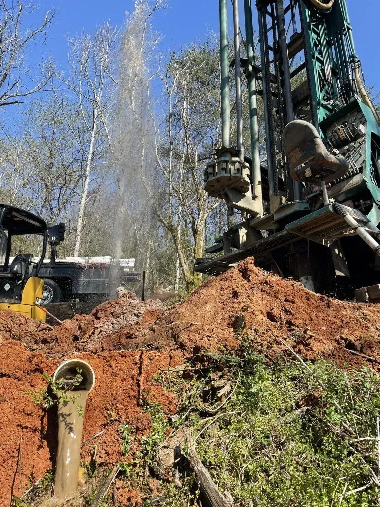 Well drilling rig spraying water, drilling into red clay, with small equipment and trees visible.