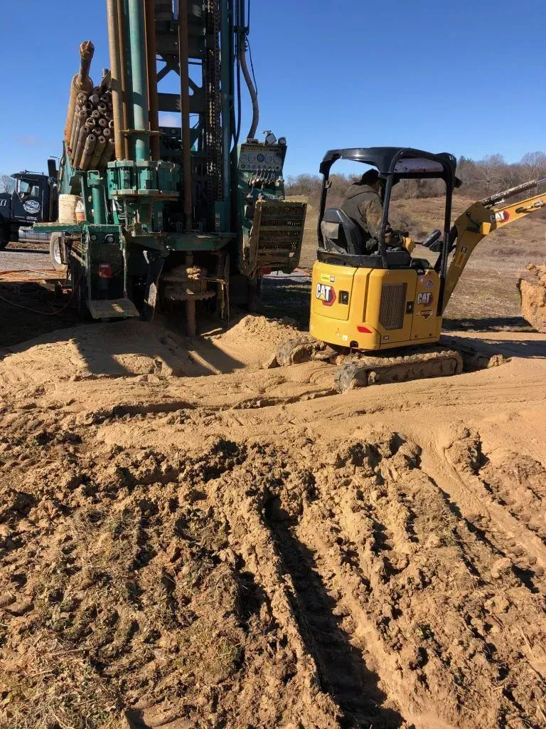 Yellow excavator and drilling rig on sandy ground.