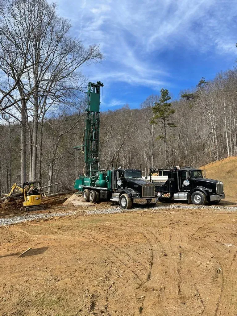 Well drilling rig and trucks on a construction site in a wooded area under a blue sky.