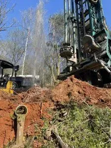 Well drilling rig spraying water, surrounded by red soil, trees, and other machinery.