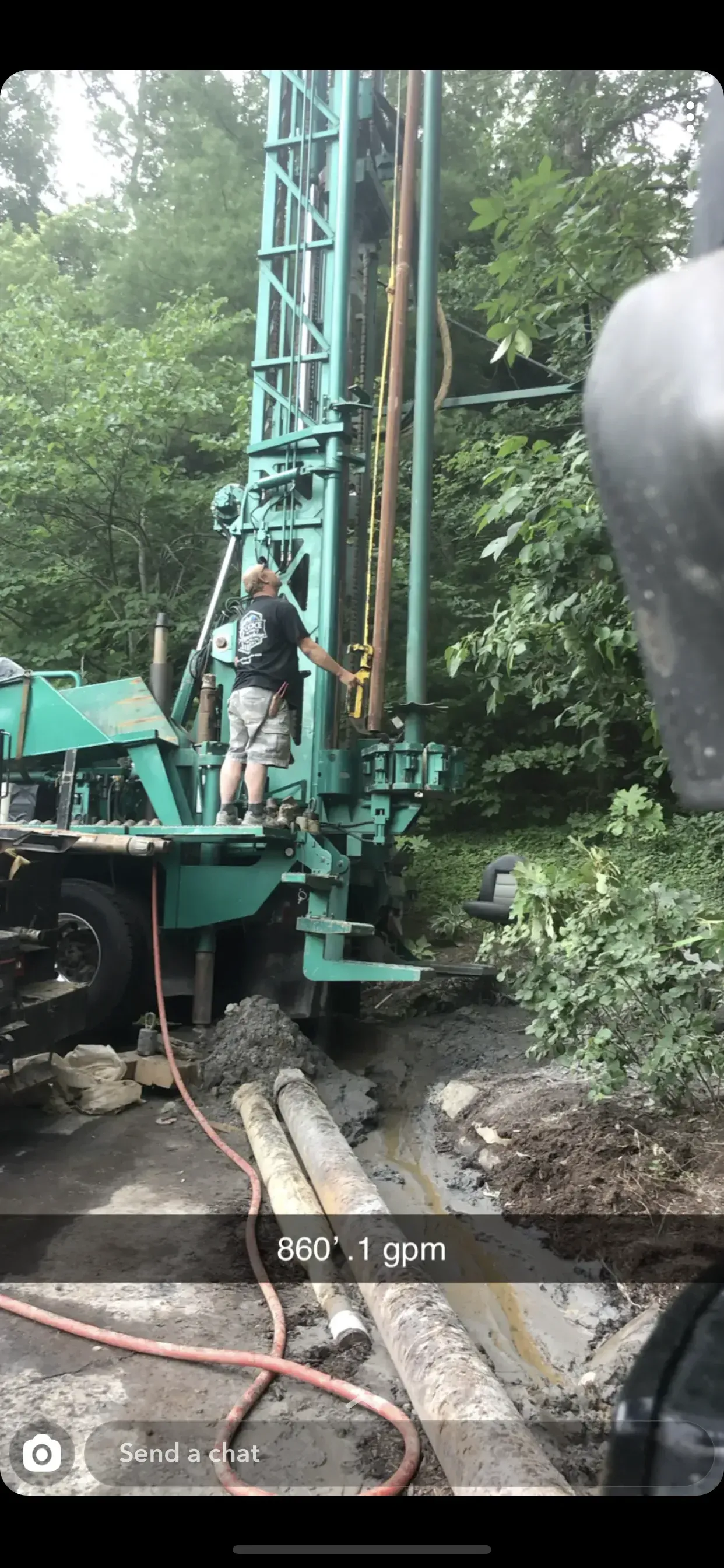 Man operating a drilling rig in a wooded area; green machinery, gray dirt, and trees.