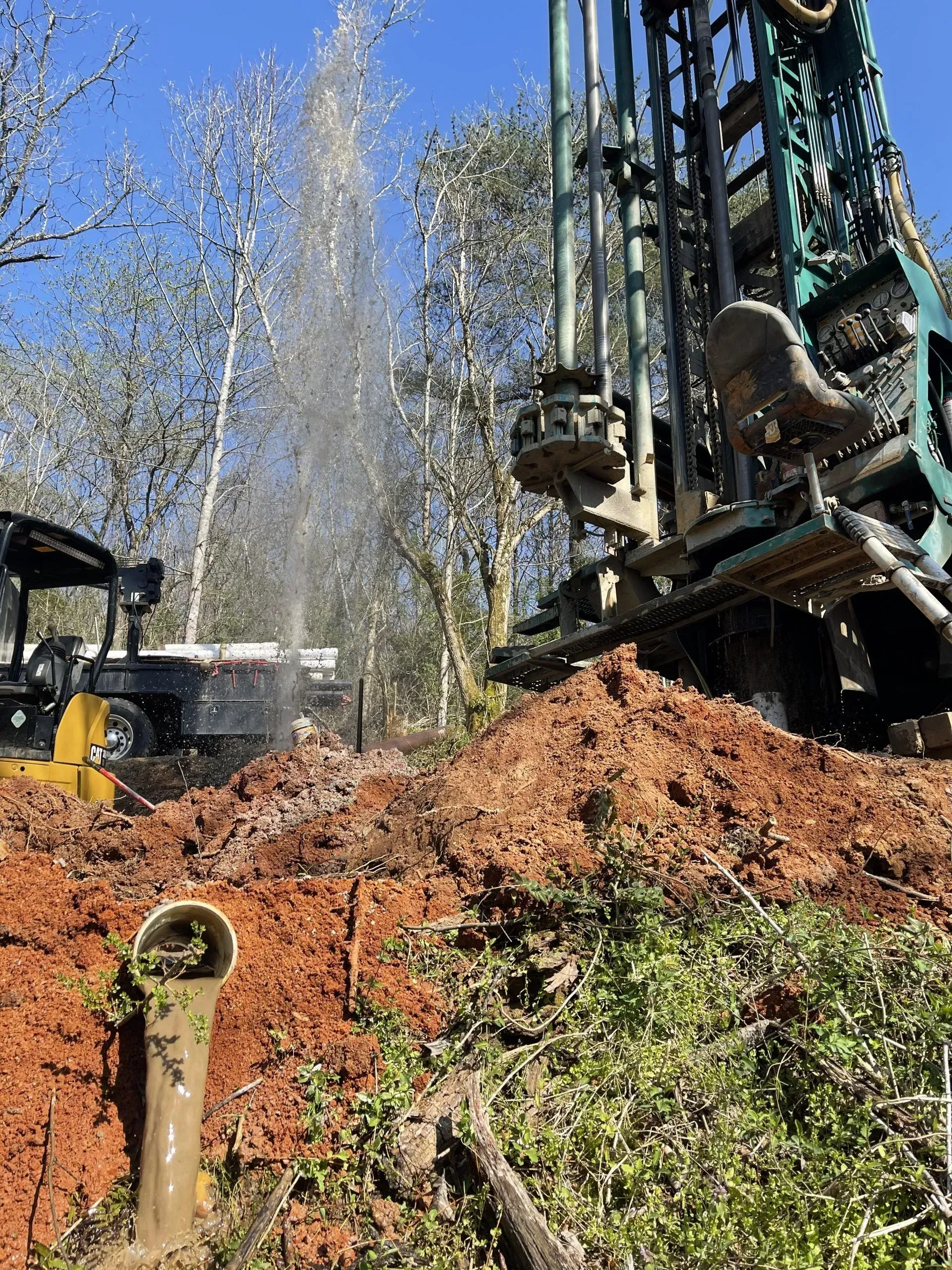 Drilling rig spurting water into the air, over a mound of dirt and greenery, with a small vehicle nearby.