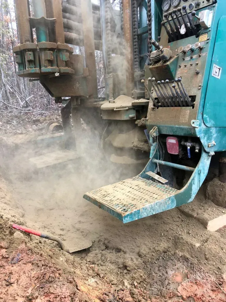 Drilling rig operating on muddy ground, kicking up dust; green and tan machinery.