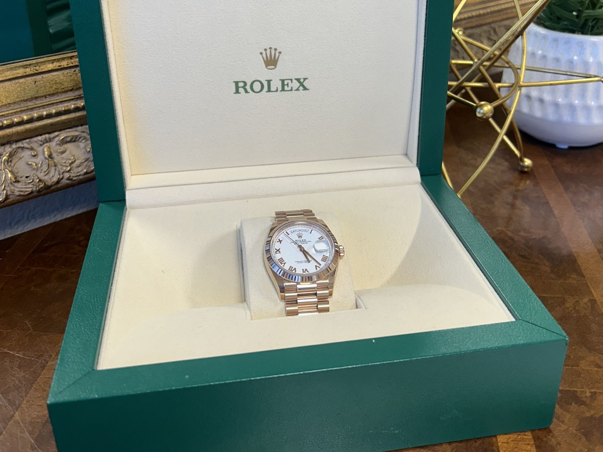 A gold Rolex Day-Date watch displayed inside an open, green-and-cream-colored luxury watch box.