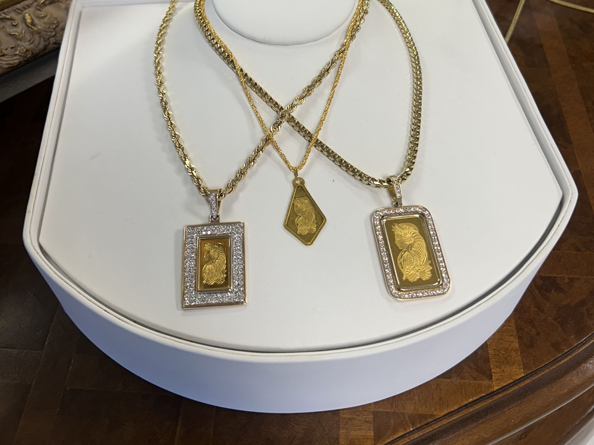 Three gold necklaces with rectangular and diamond-shaped gold pendants are displayed on a white stand.