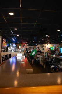 Bar interior with customers and staff. Long bar, dark ceiling, and overhead lighting.