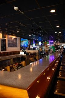 Bar interior with a long wooden bar, stools, and dark ceiling with lights.