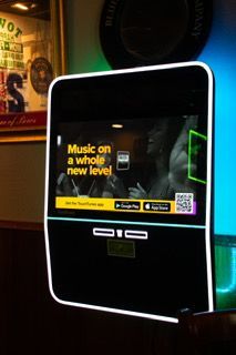 Jukebox with a screen showing an advertisement for a music app. It is black with a white outline and is mounted on a wall.