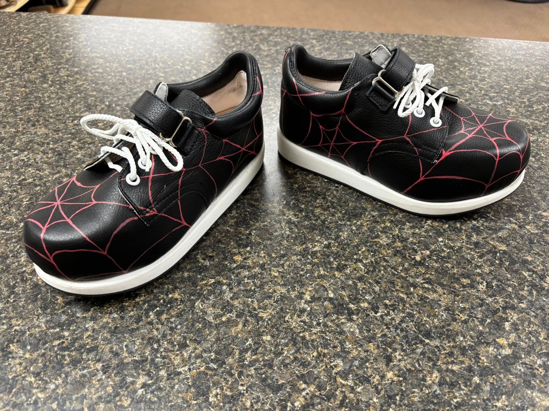 Black shoes with spiderweb design and white soles, on a textured surface.
