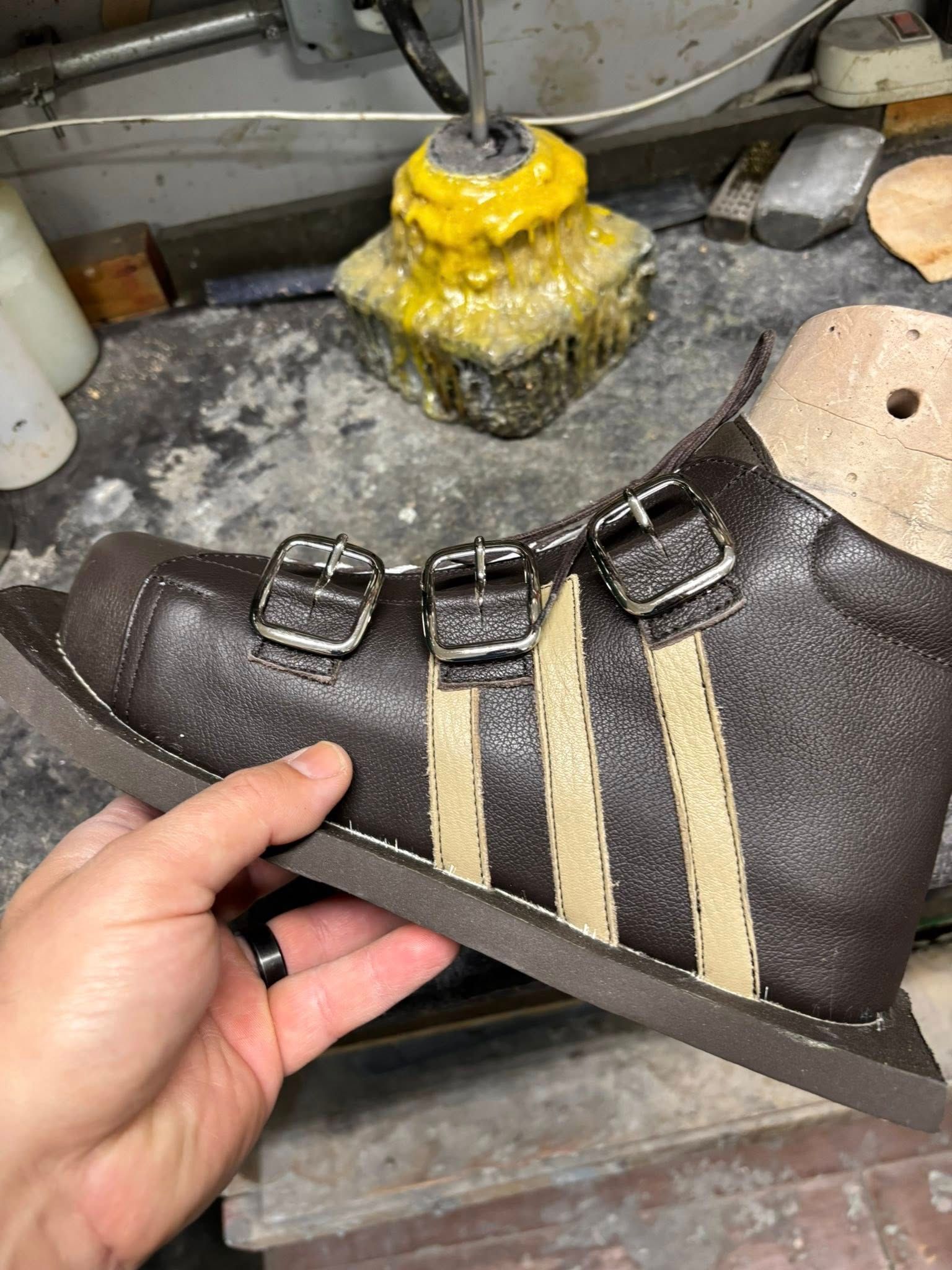 A person holding a brown, custom-made shoe with beige stripes and buckles, in a workshop setting.