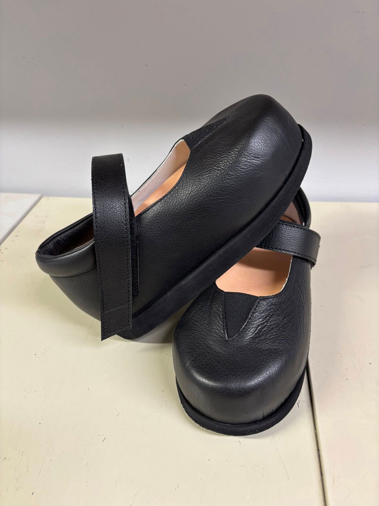 Black leather Mary Jane style shoes with a strap and round toe, stacked on a white surface.