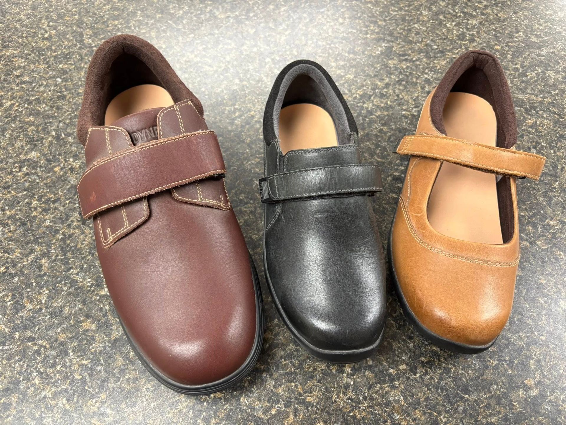 Three leather shoes of different colors (brown, black, tan) with velcro straps, on a dark countertop.