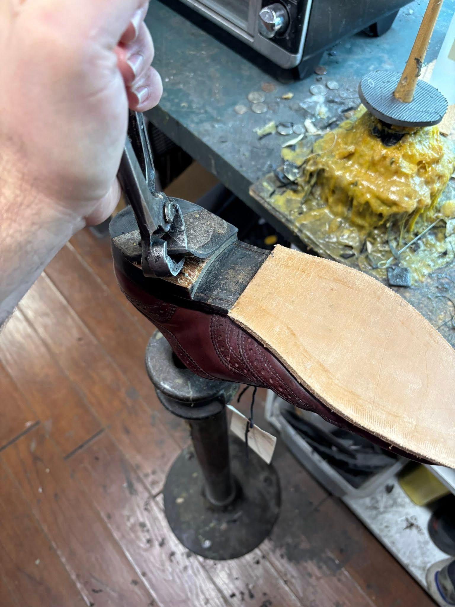 Person using pliers to work on the sole of a shoe, near a workbench with wax and a toaster oven.