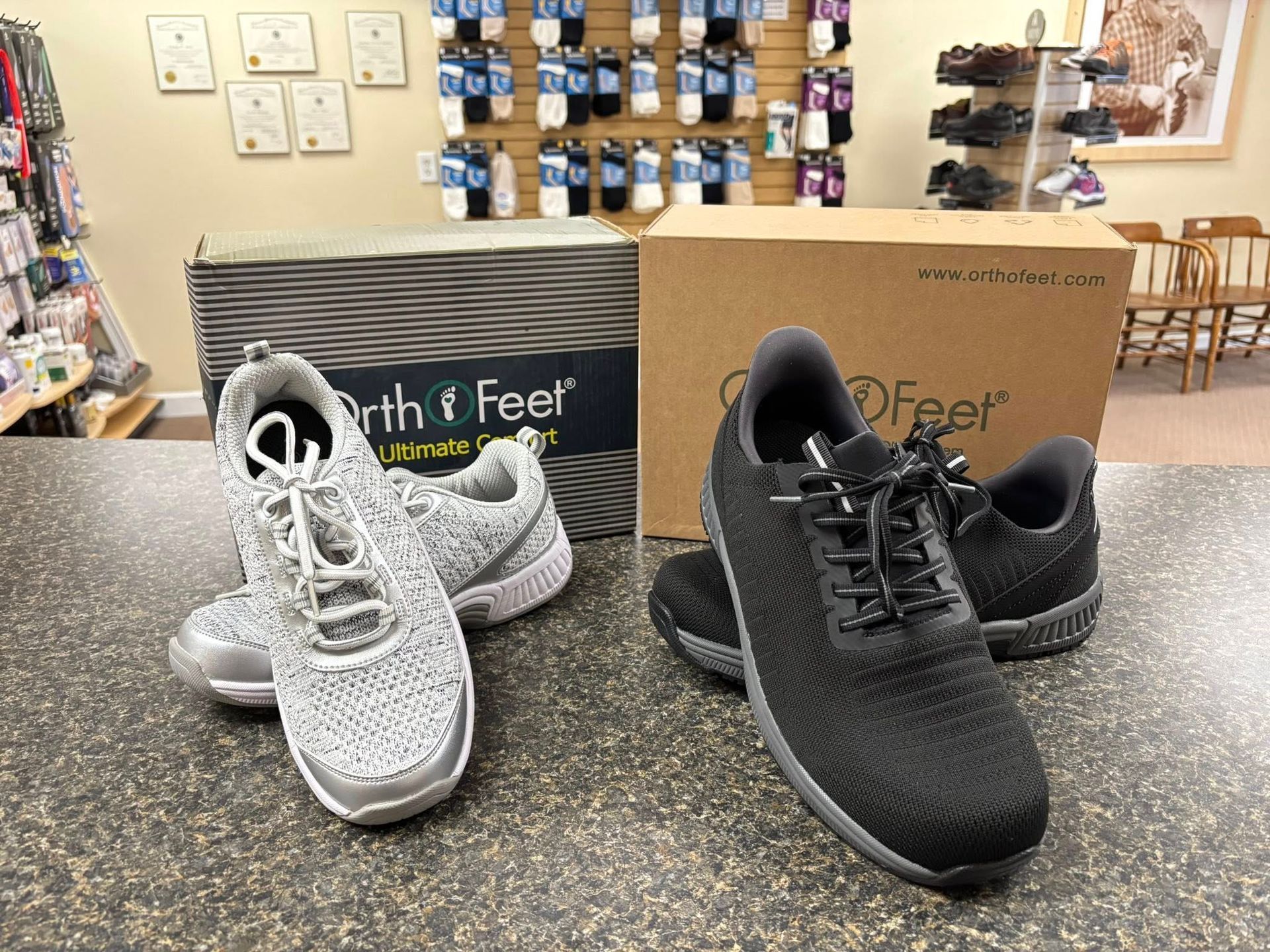 Two pairs of orthotic sneakers, grey and black, on a counter in a shoe store.
