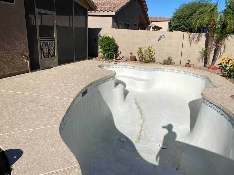A dry, light-colored swimming pool with a textured stone deck in a residential backyard with a block wall fence.