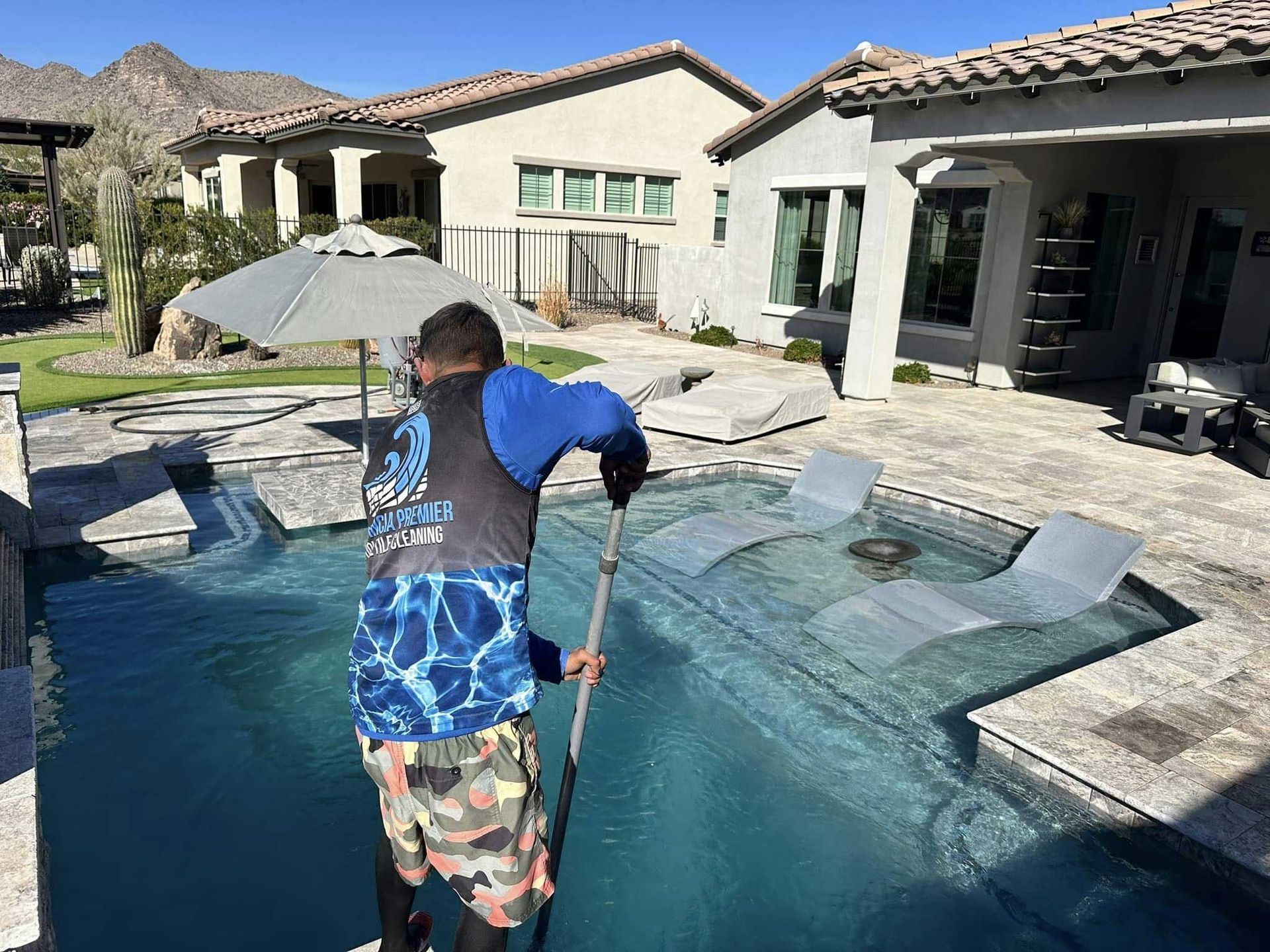 A technician in a blue shirt cleans a backyard swimming pool with a pole in a desert-landscape setting.
