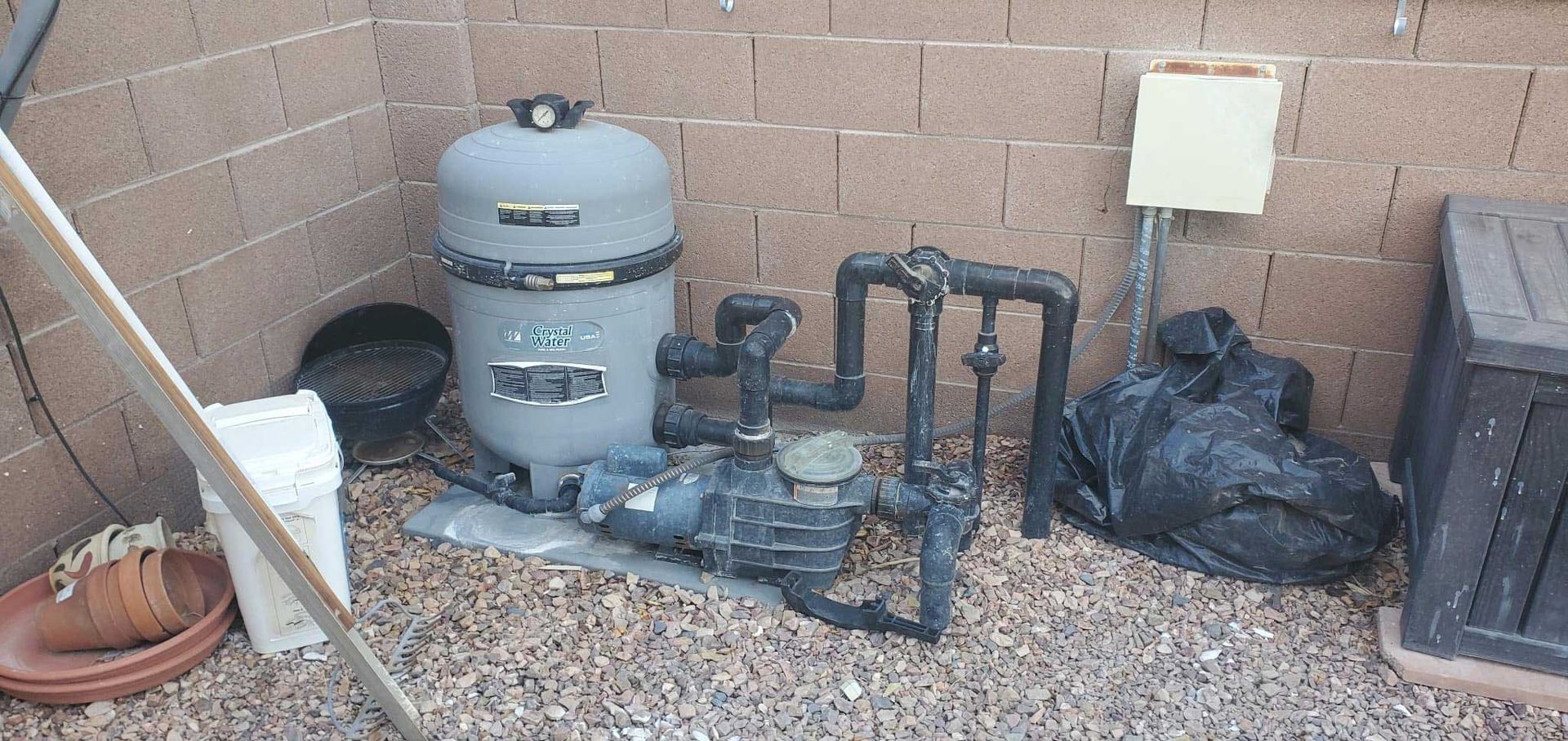 A grey pool pump and filtration tank system positioned against a cinder block wall on a gravel surface.