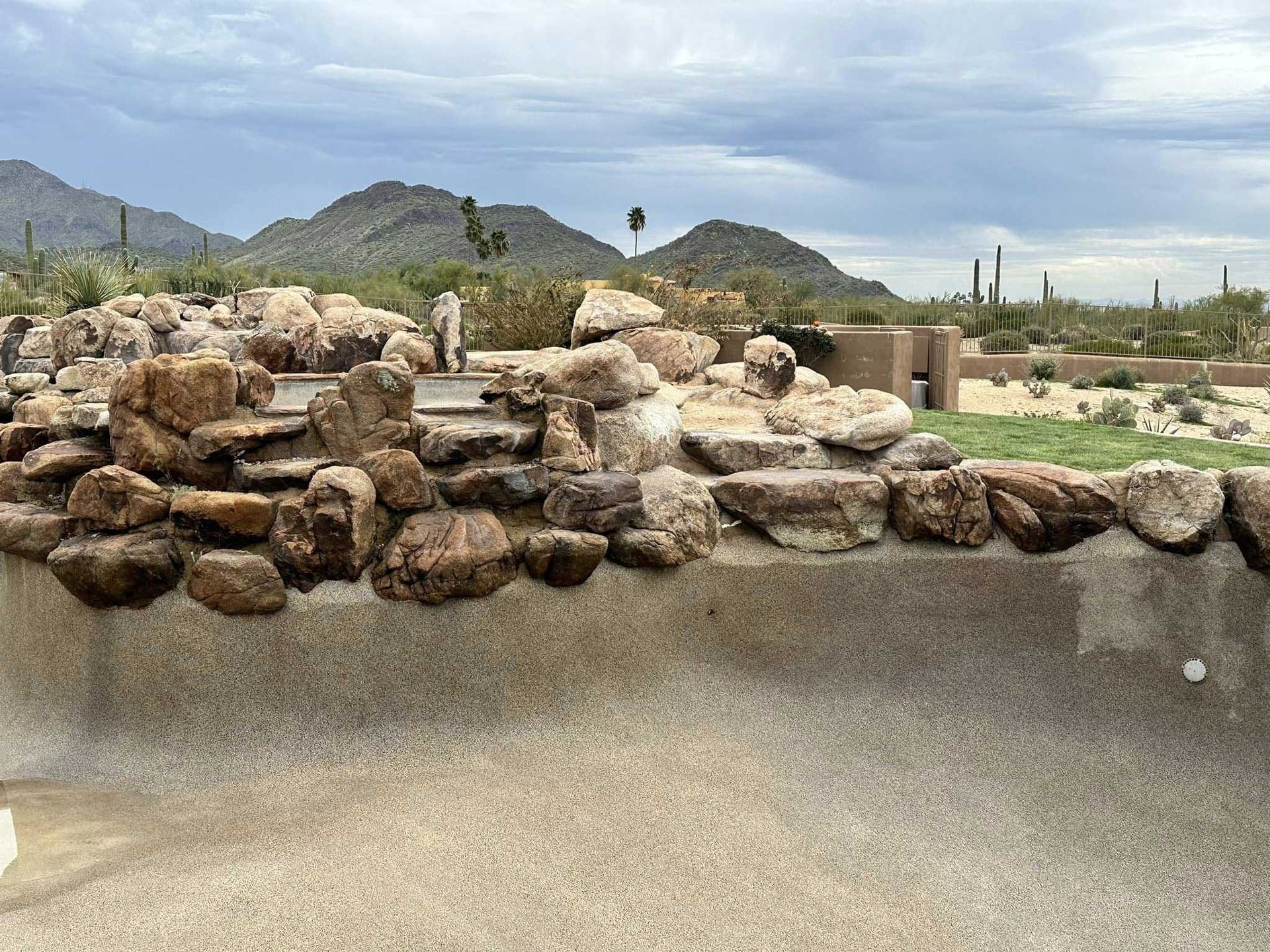 A rocky waterfall feature bordering a dry, pebble-textured swimming pool with mountains and desert flora in the distance.