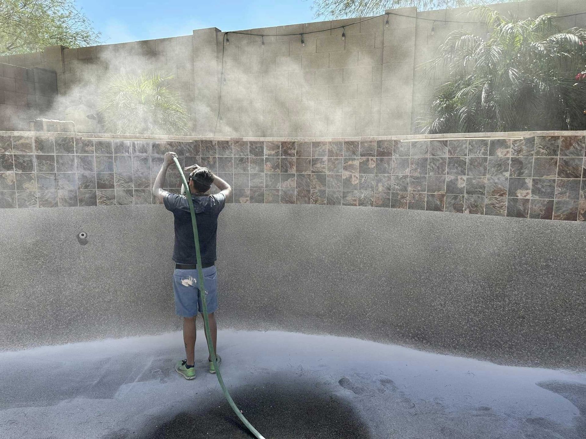 A person stands in an empty pool, pressure-washing a tiled wall, creating a fine mist of spray.