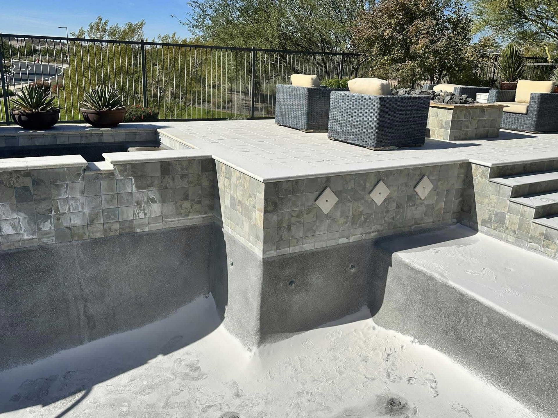 A concrete pool deck under construction with stone tiling, steps, and outdoor furniture on a patio above.