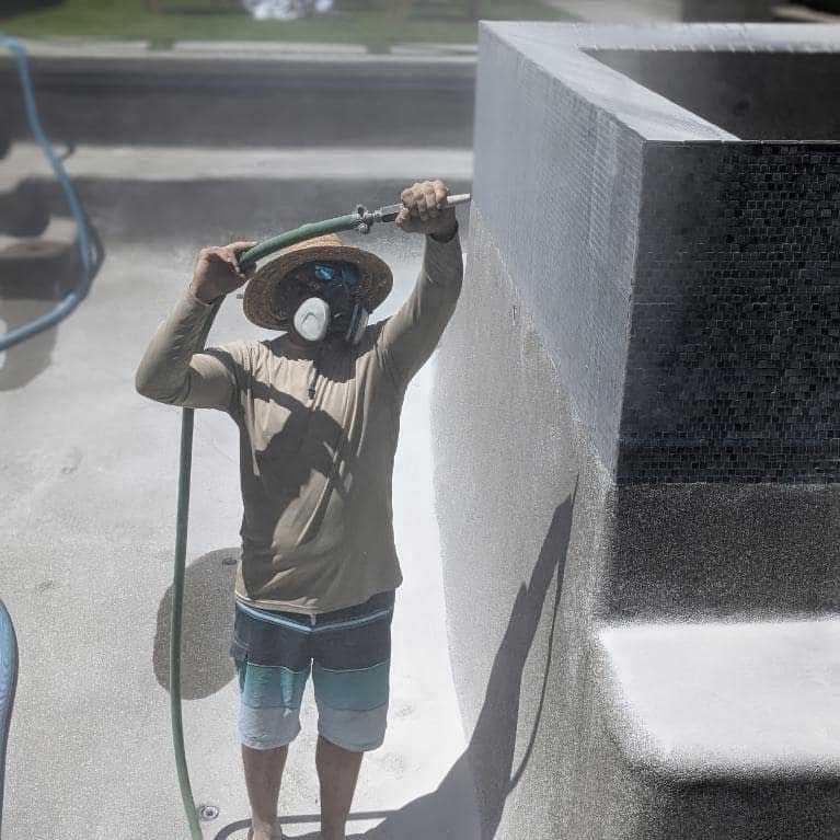A worker wearing a hat and respirator sprays a textured pool surface with a hose.