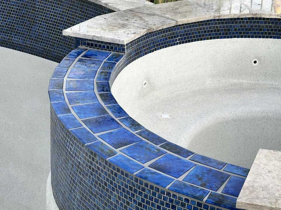 A curved pool spa featuring deep blue square tiles, a light stone coping edge, and a smooth, light-colored plaster basin.
