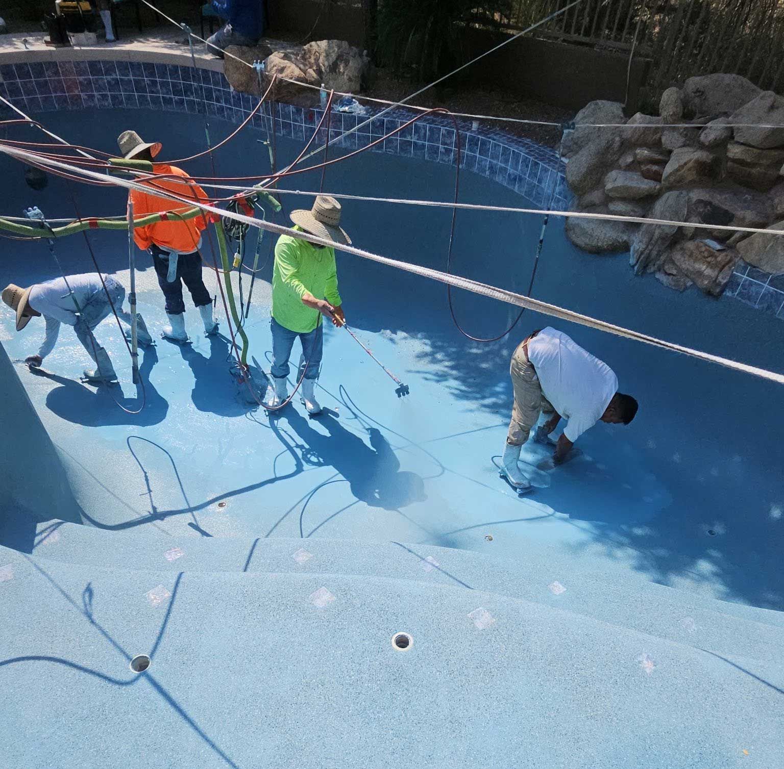 Workers in safety gear applying a light blue finish to the interior of a swimming pool.