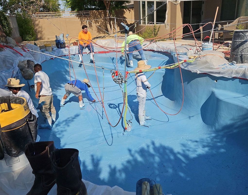 Workers in white protective gear spray-coat the interior of a blue swimming pool, using ropes and hoses to reach surfaces.