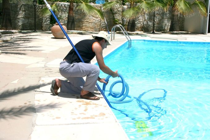 A person kneeling on a pool deck, using a long pole and hose to clean a bright blue swimming pool.