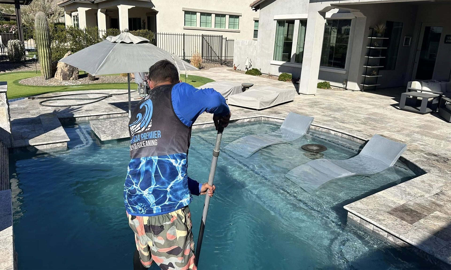 A worker uses a long-handled tool to clean a swimming pool with lounge chairs on a tanning ledge.
