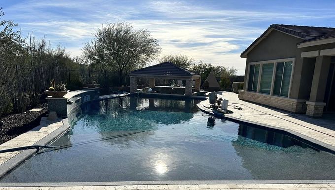 A swimming pool with a covered patio and house in the background under a sunny blue sky.