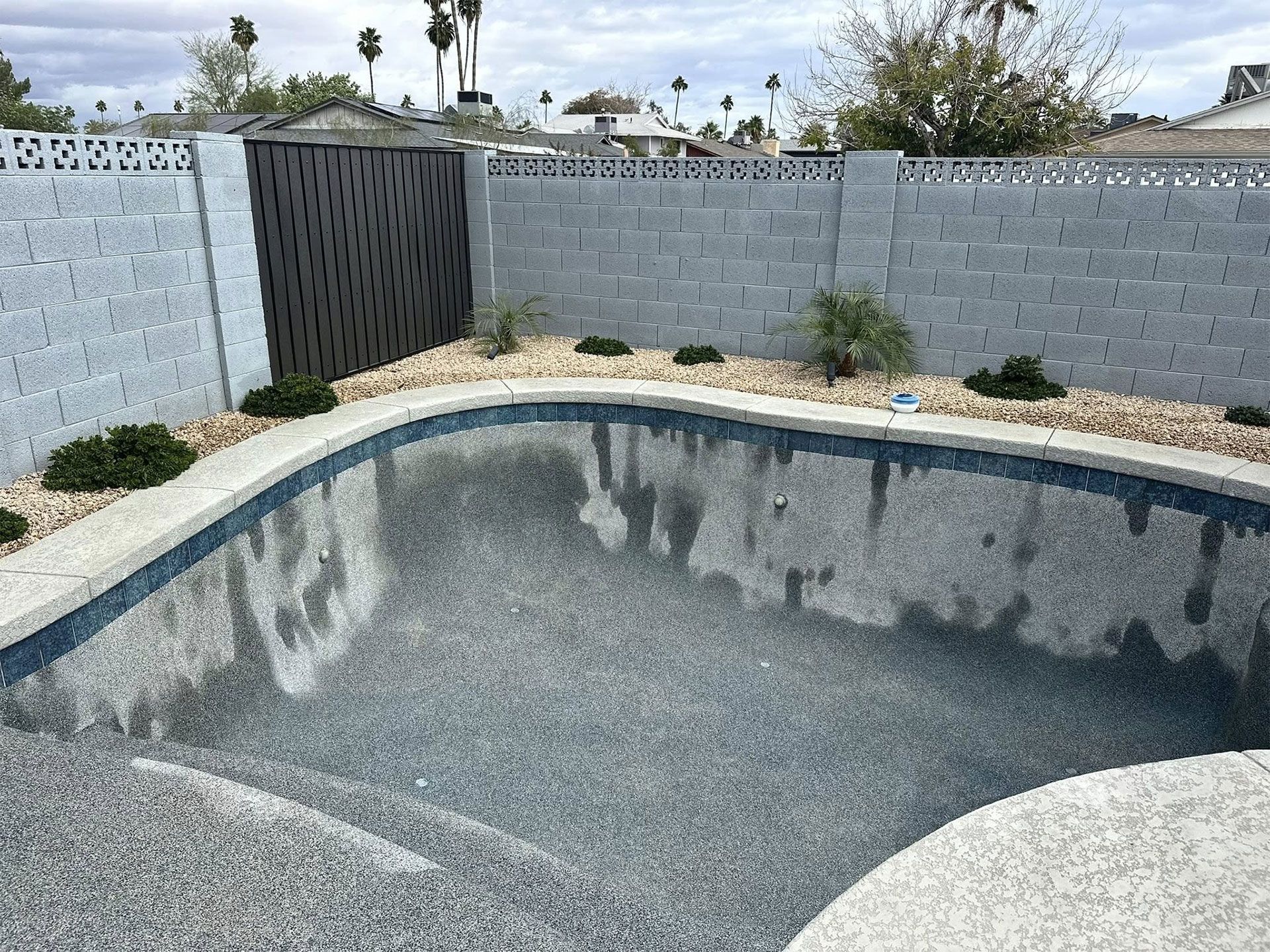 An empty residential swimming pool with grey pebble-finish walls, surrounded by decorative rocks and a concrete wall.
