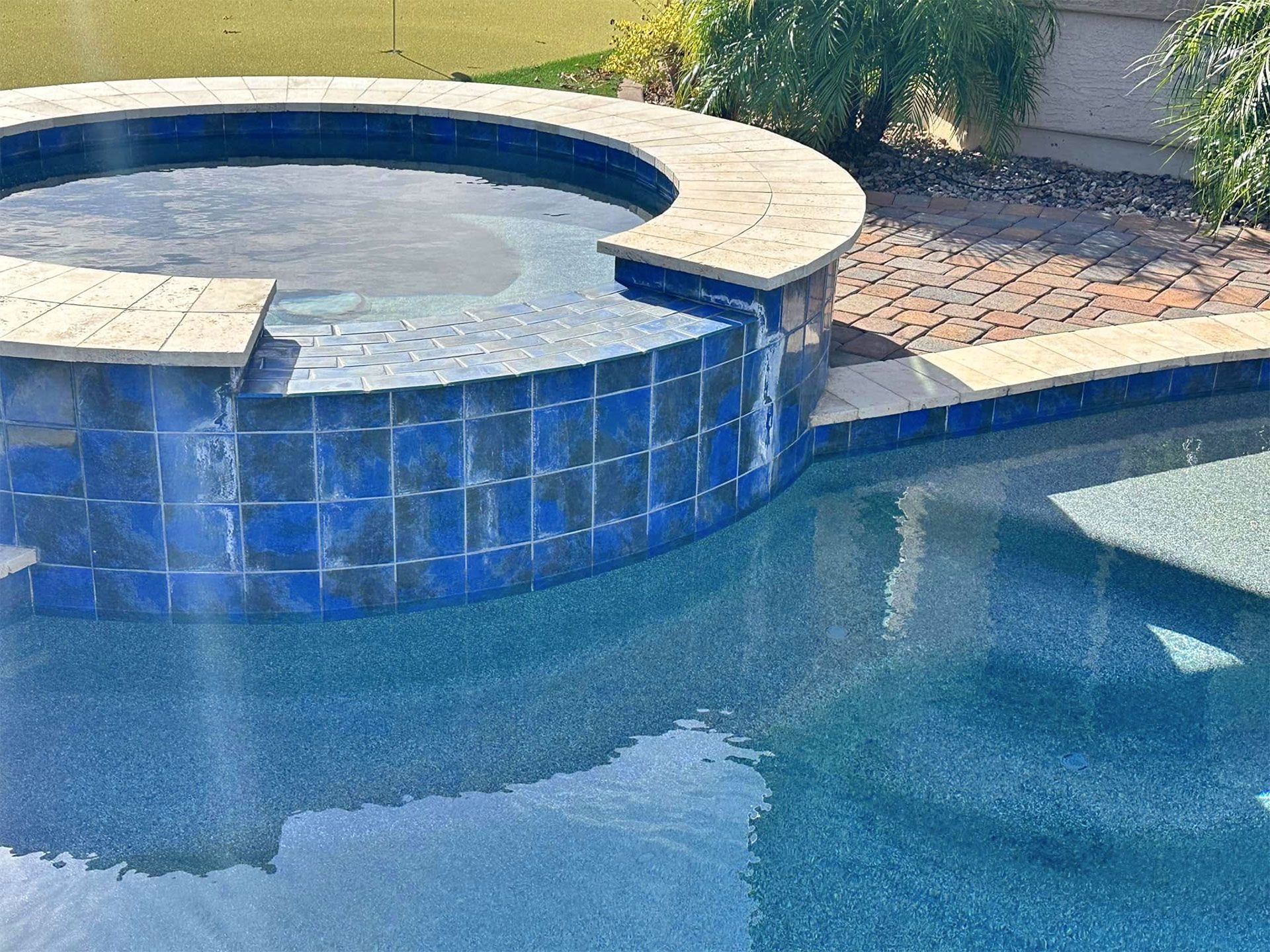 A circular hot tub with blue tiled walls overflowing into a swimming pool with a stone patio and landscaping nearby.