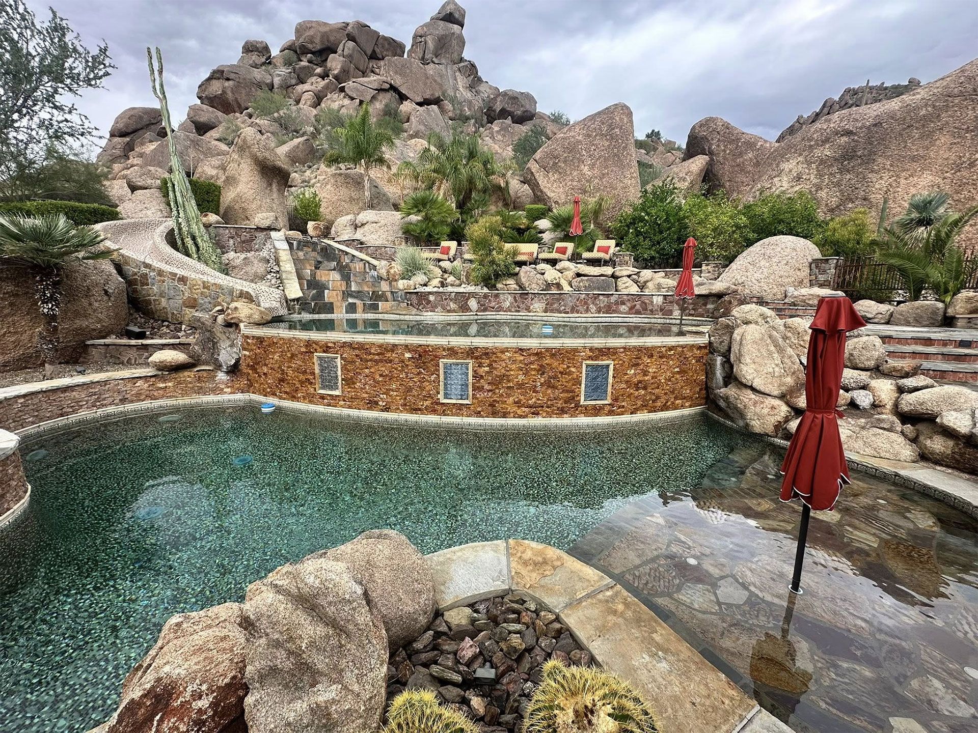 A multi-level pool with a stone wall and built-in lighting, nestled against a rocky desert hillside with a red umbrella.