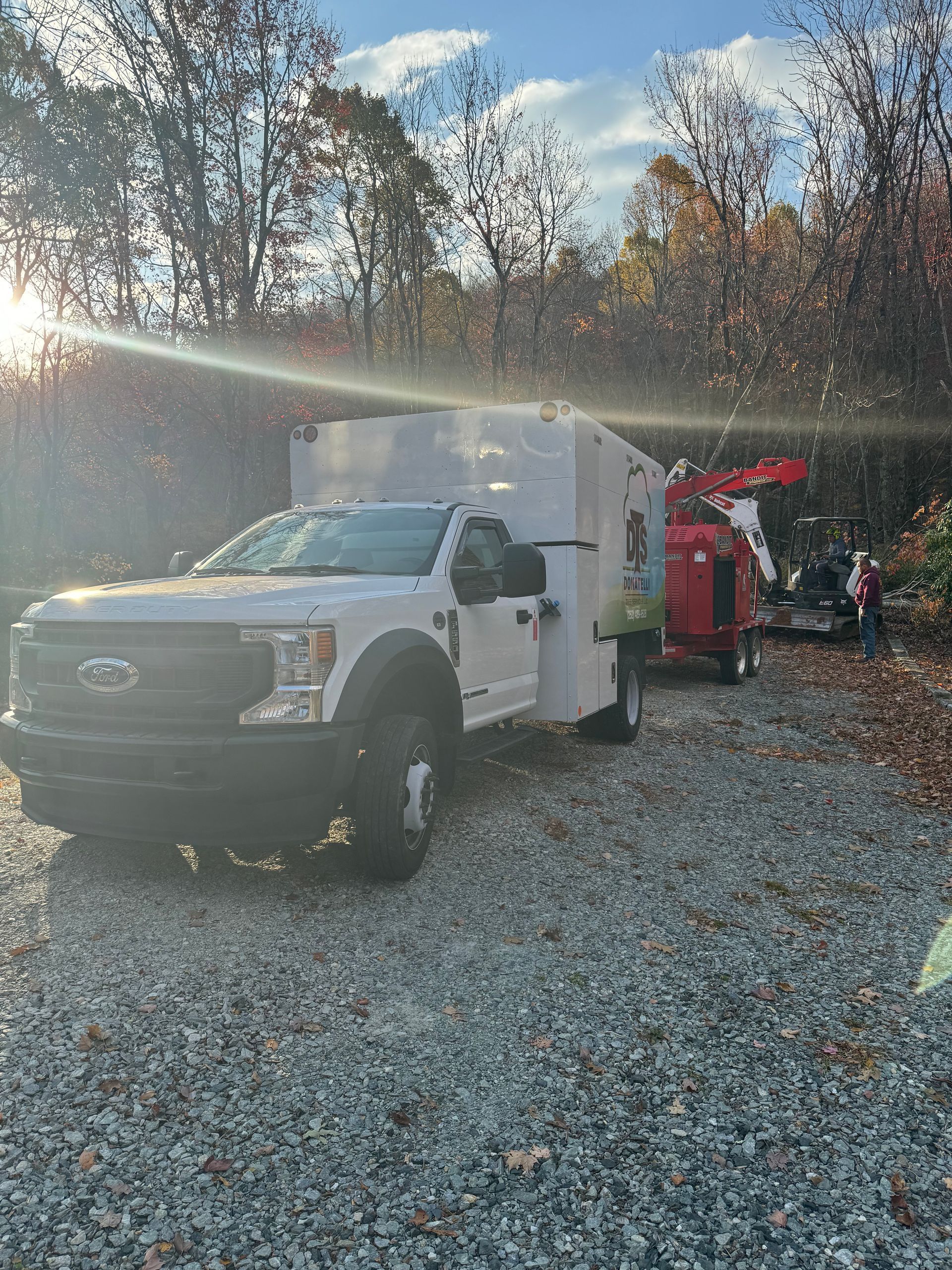 White work truck and wood chipper in a wooded area with sunlight. People visible in the background.
