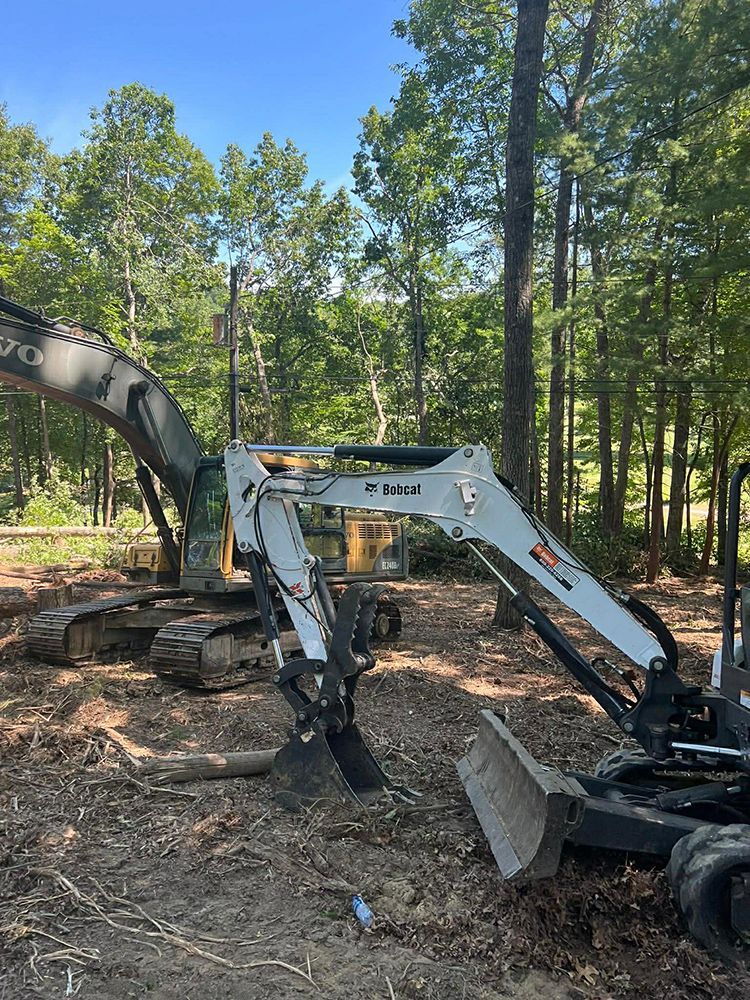 A white Bobcat excavator stands in the foreground, with a larger yellow Volvo excavator in the background in a wooded area.