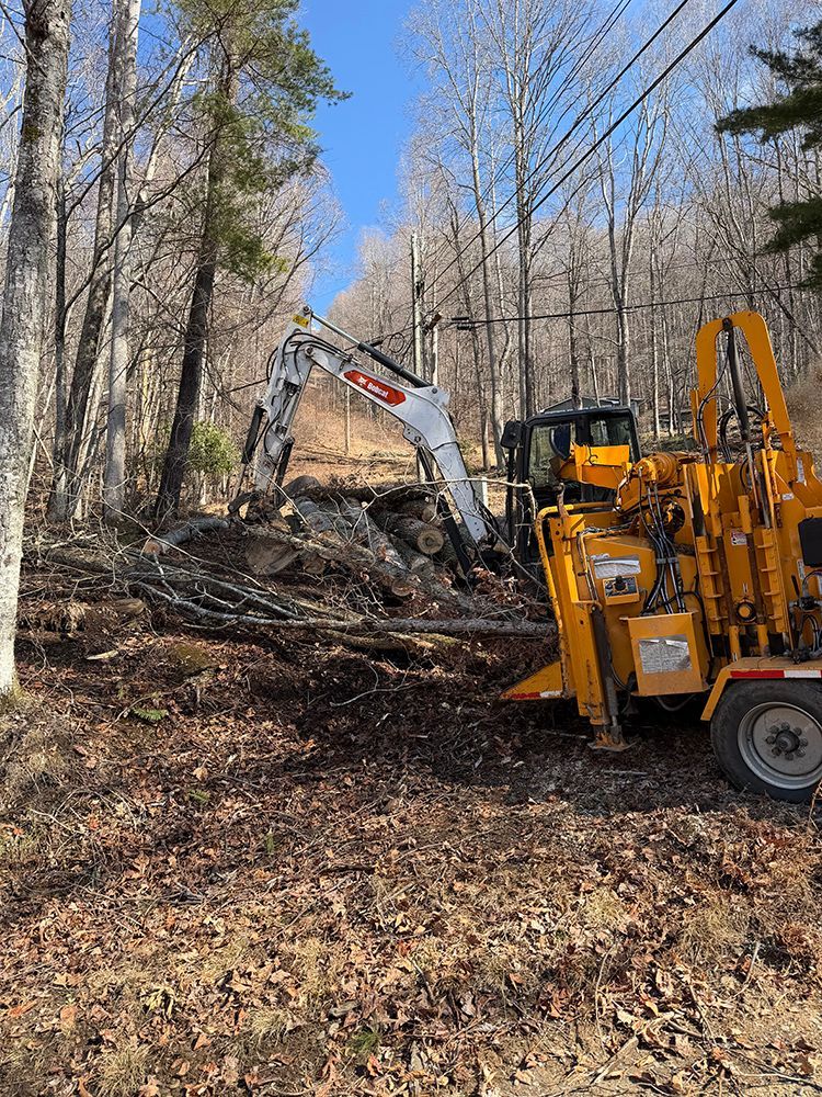 A yellow utility truck and a white Bobcat excavator working on a wooded hillside under a clear blue sky.