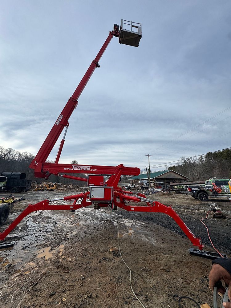 A red spider lift with its outriggers extended, parked on a muddy, outdoor work site under a cloudy sky.