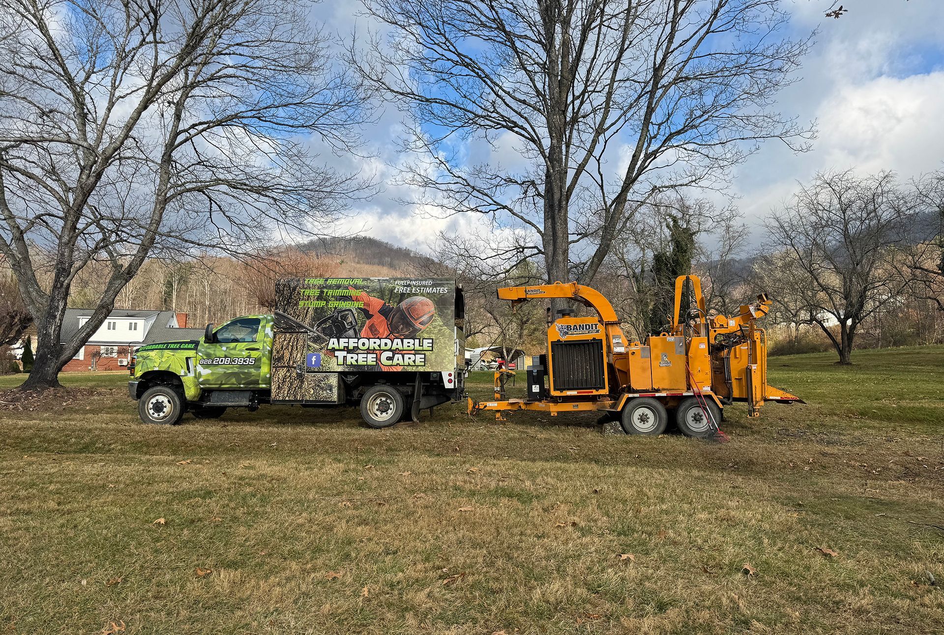 A green utility truck with a camo-patterned bed towing a large, yellow wood chipper on a grassy field.