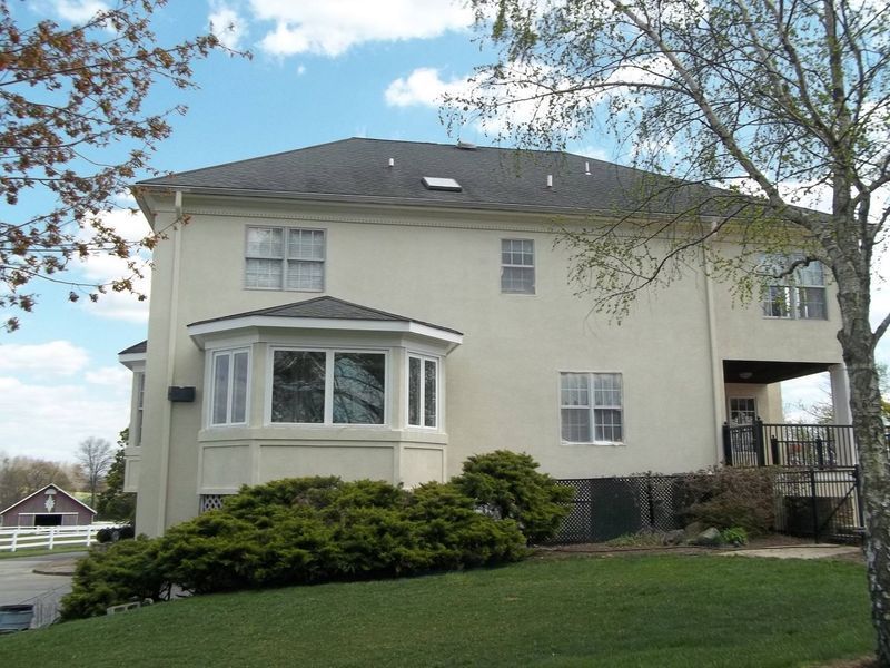 Beige two-story house with a bay window, deck, and dark roof. Green lawn and a small red barn are in view.