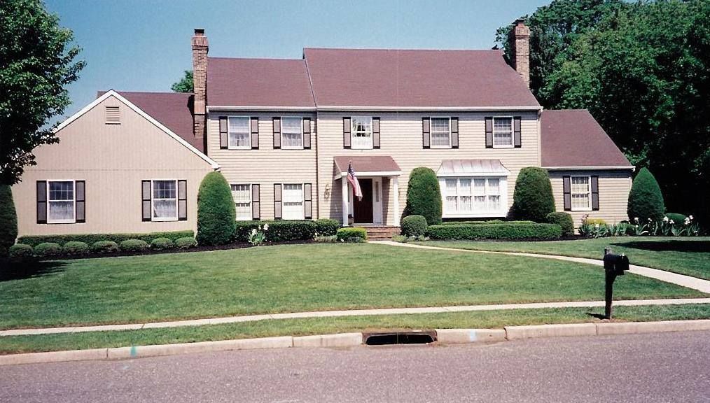 Tan two-story house with brown roof and black shutters. Green lawn, trees, and street in front.