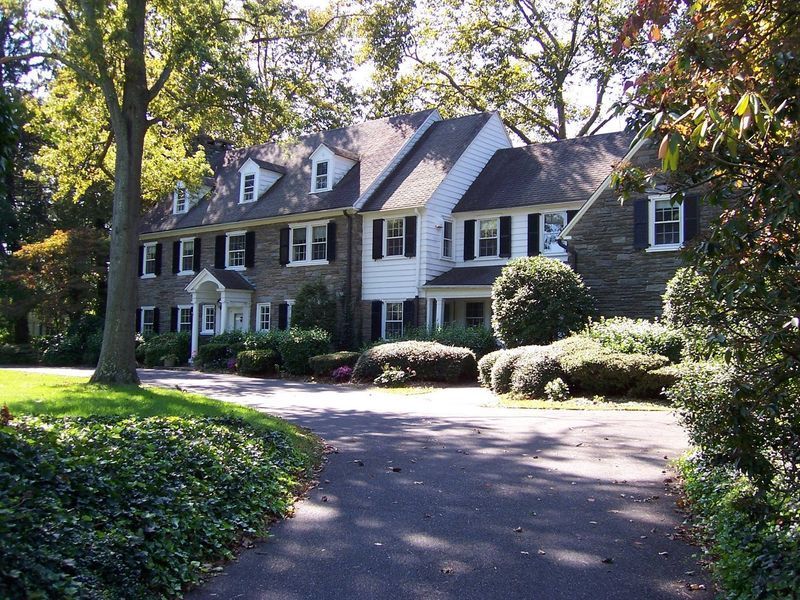 Large, two-story stone house with a long driveway. White trim, multiple windows, and dormers. Green lawn and trees.