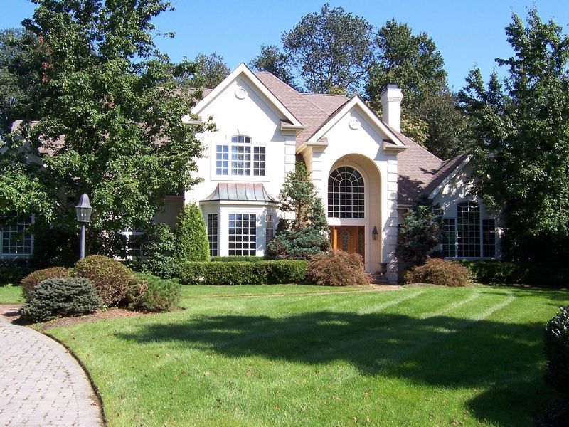 Two-story beige house with arched doorway and multiple windows, surrounded by green lawn and trees under a blue sky.