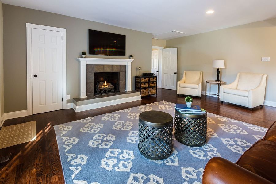 Living room with fireplace, white chairs, and patterned blue rug. Dark hardwood floors and tan walls.