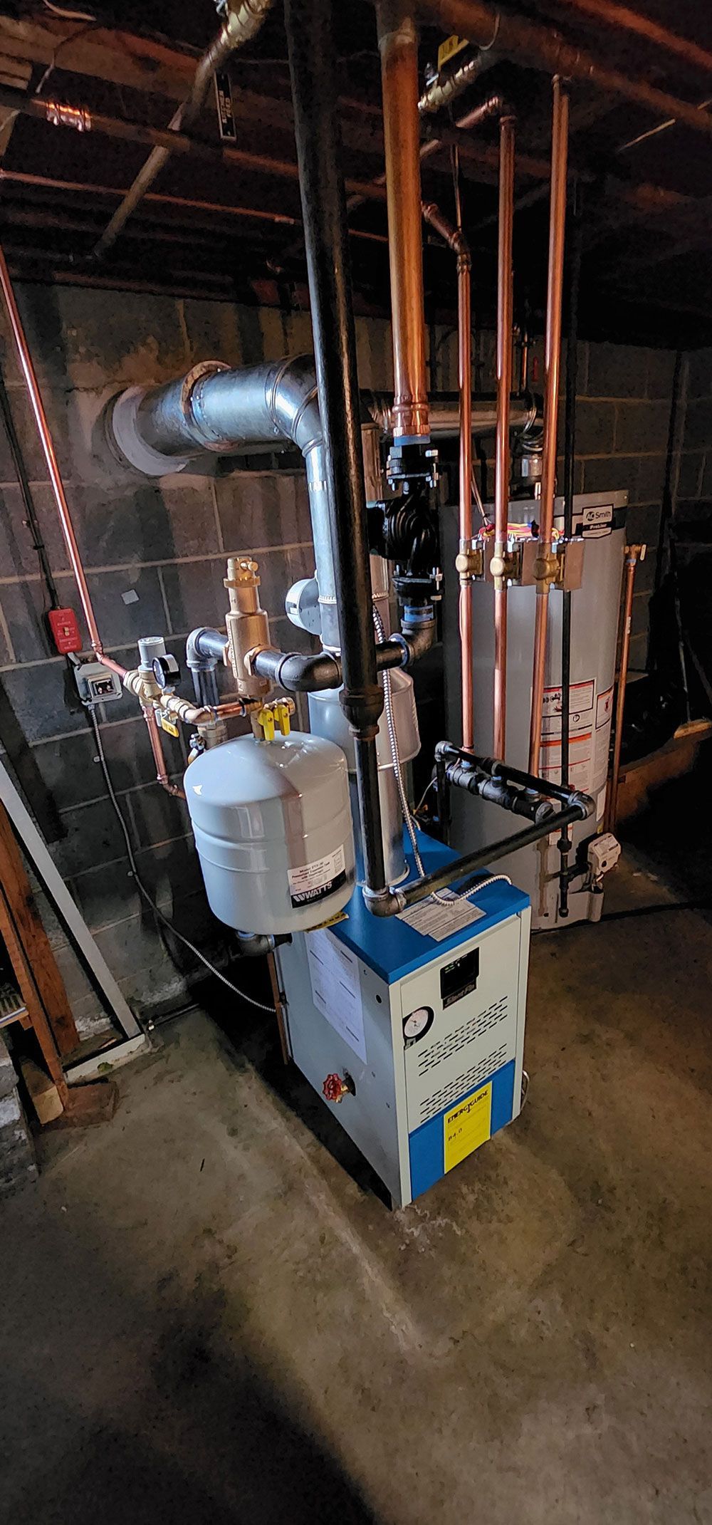 A blue and white boiler is sitting in a basement surrounded by pipes.